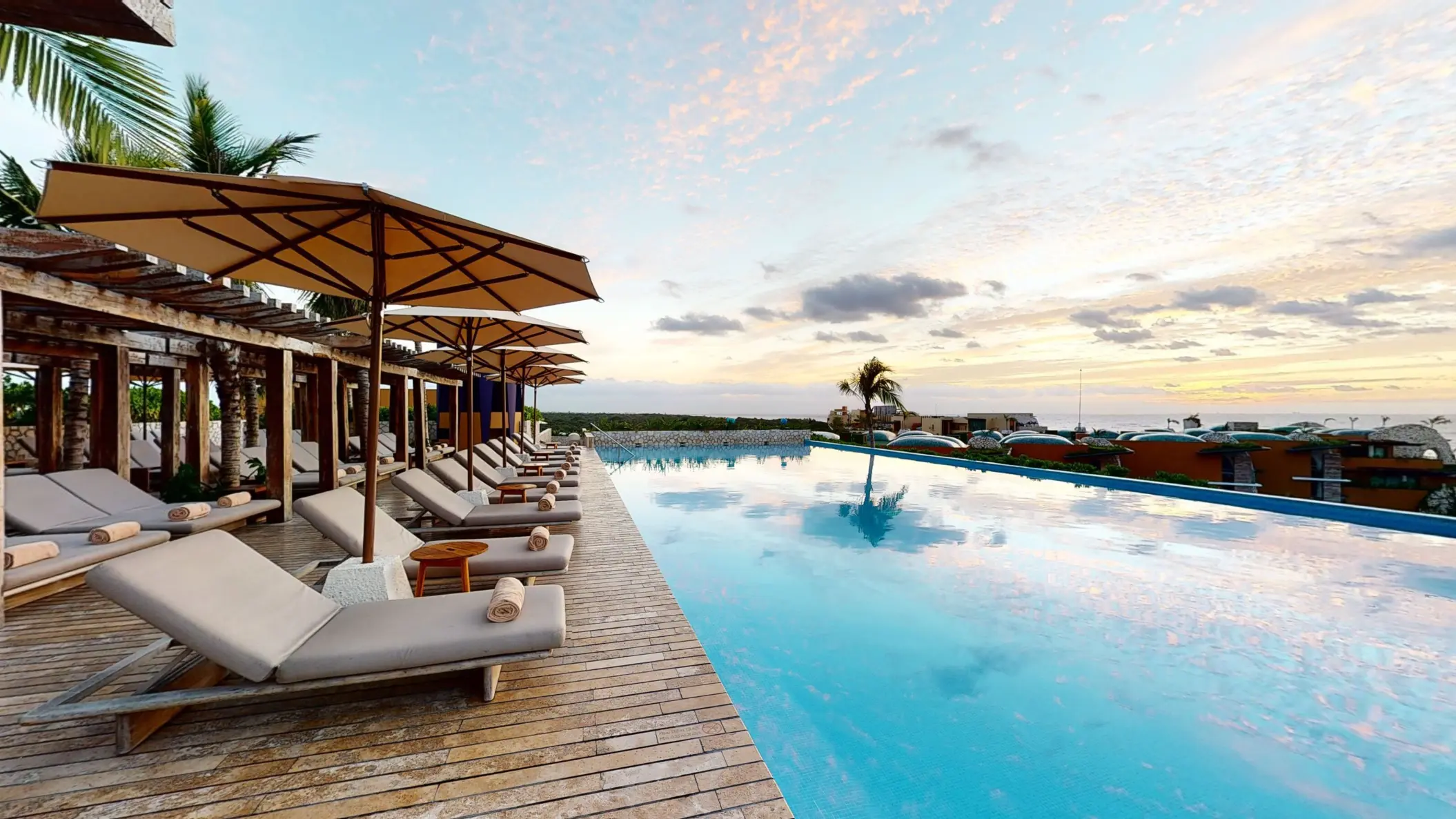 Infinity-edge pool with lounge chairs and umbrellas, overlooking the sea and tropical rooftops at sunset.