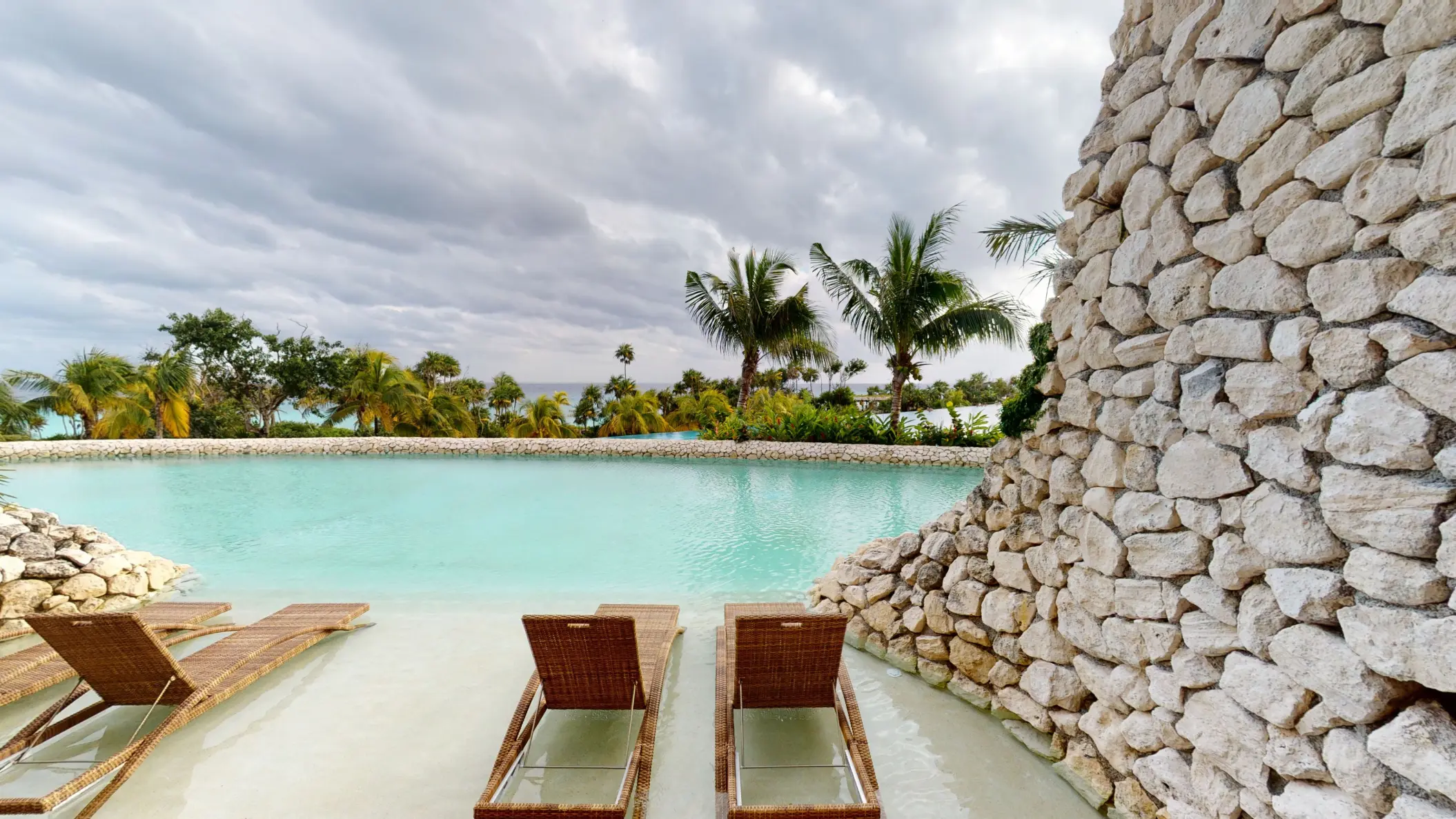 Lagoon-style pool with submerged wicker loungers and a stone wall, surrounded by palm trees.