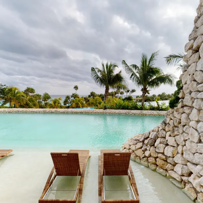 Lagoon-style pool with submerged wicker loungers and a stone wall, surrounded by palm trees.