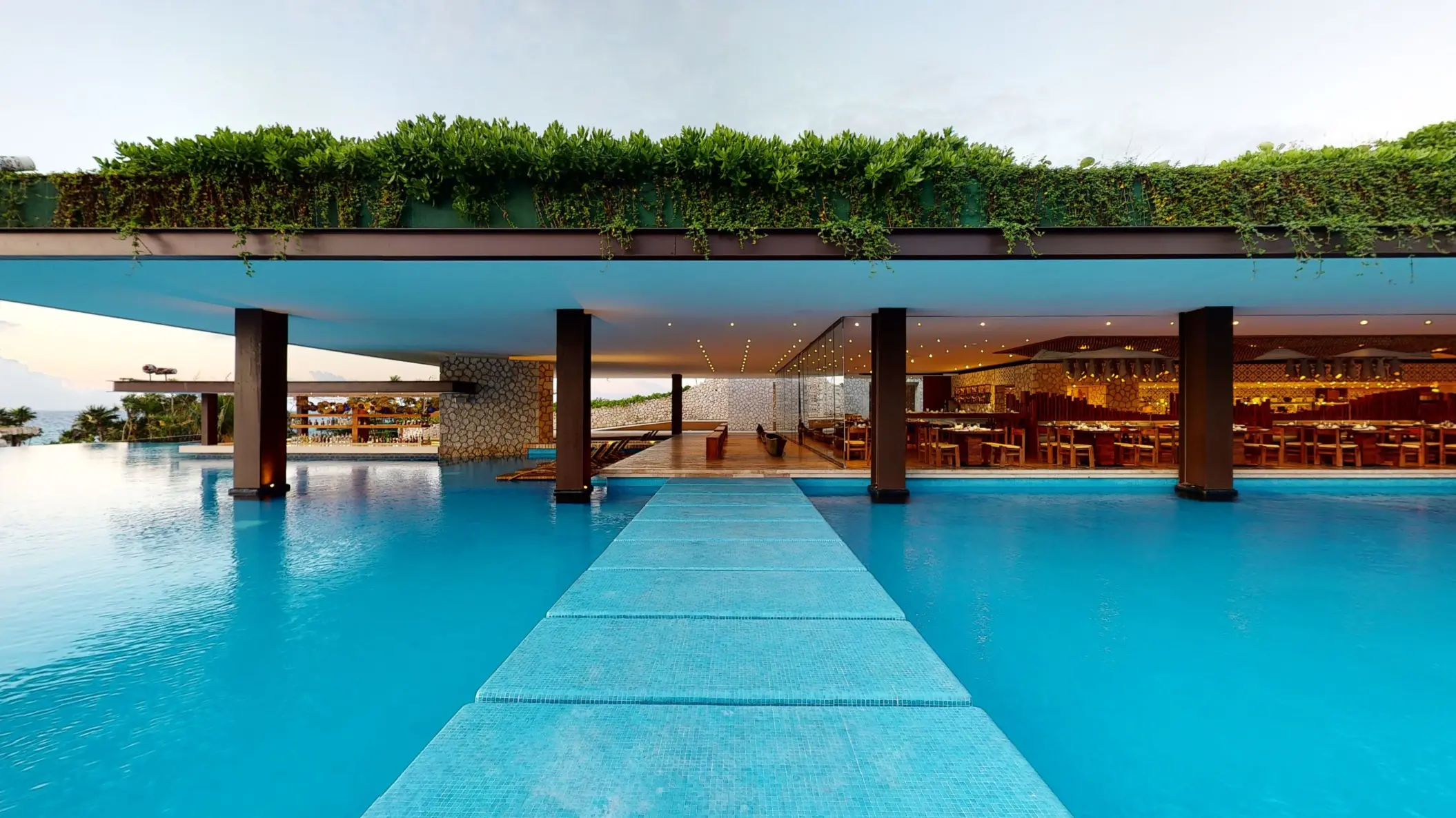Walkway across infinity pool leading to open-air restaurant with lush rooftop garden.