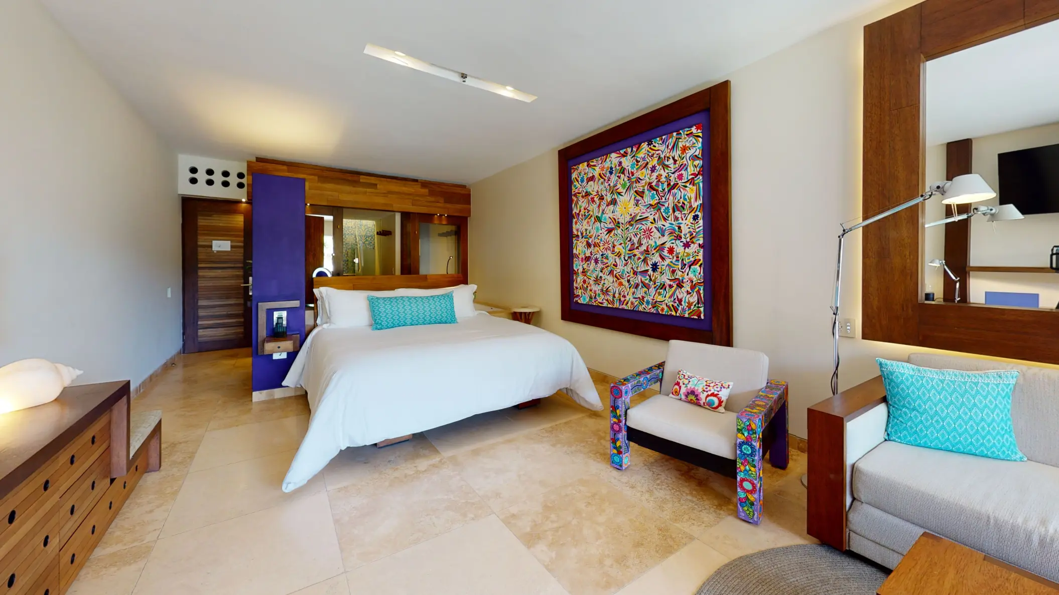 Modern guest room with king bed, colorful Otomi-inspired artwork, and cozy seating area with turquoise pillows.