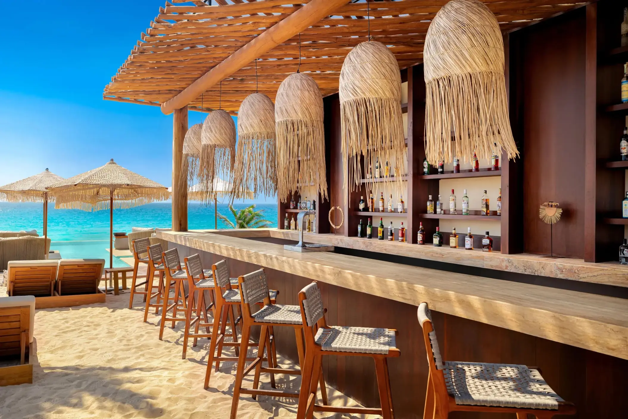 Beach bar with wooden stools, straw lamps, and ocean views under a rustic palapa-style pergola.