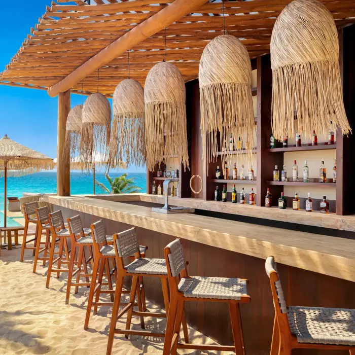 Beach bar with wooden stools, straw lamps, and ocean views under a rustic palapa-style pergola.