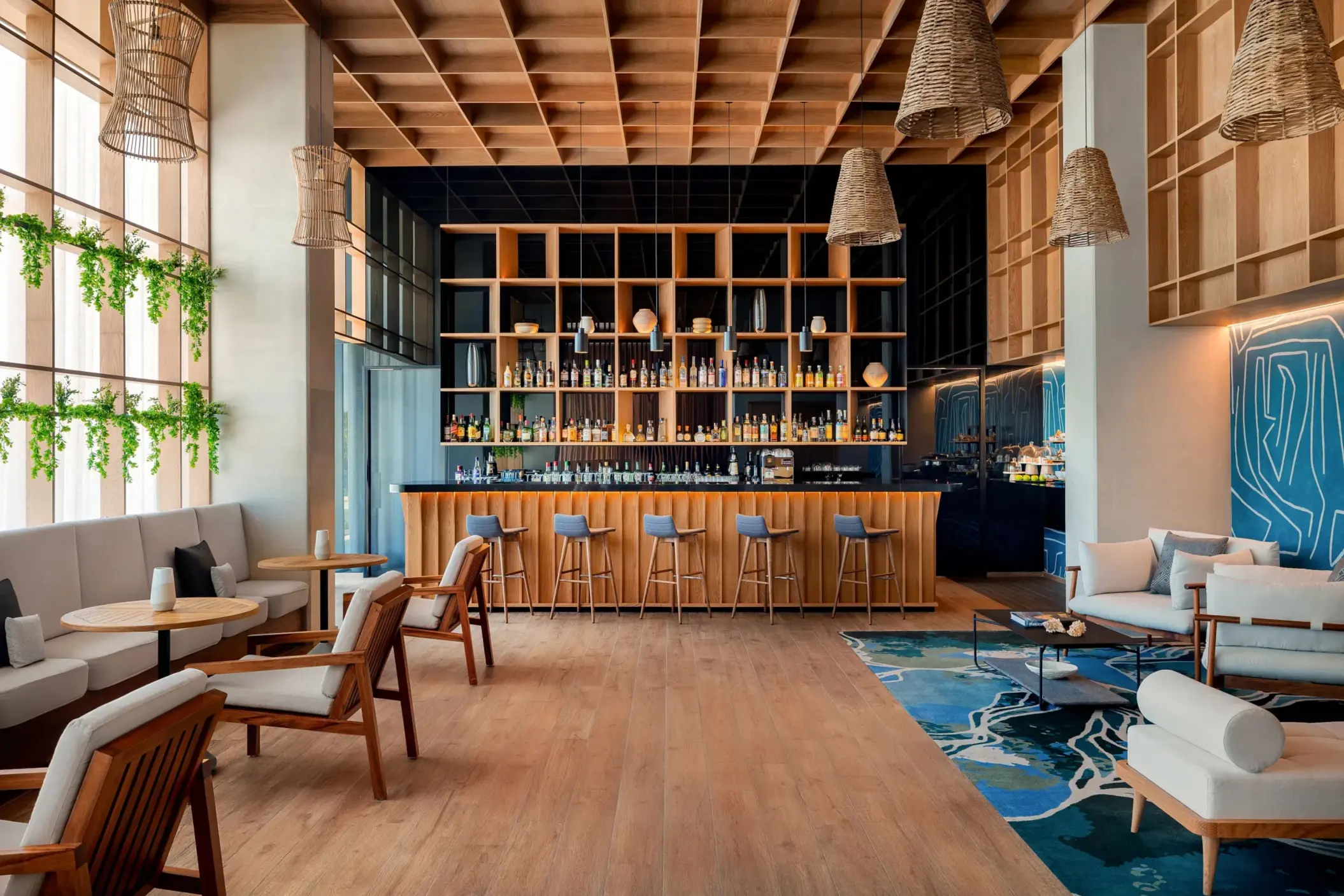 Upscale indoor lounge bar with wooden ceiling, stylish furniture, greenery, and floor-to-ceiling bottle display.