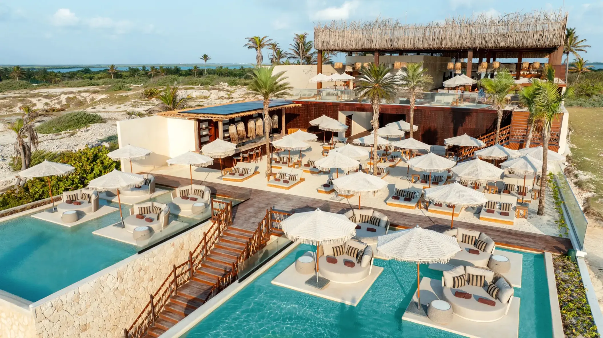 Aerial view of beach club with cabanas, palm trees, two-tiered pools, and private shaded lounge areas.