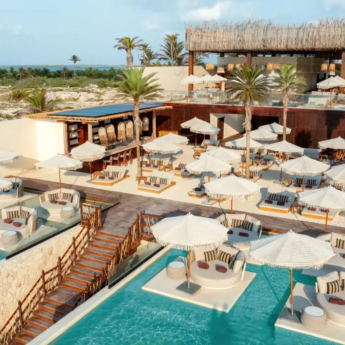 Aerial view of beach club with cabanas, palm trees, two-tiered pools, and private shaded lounge areas.