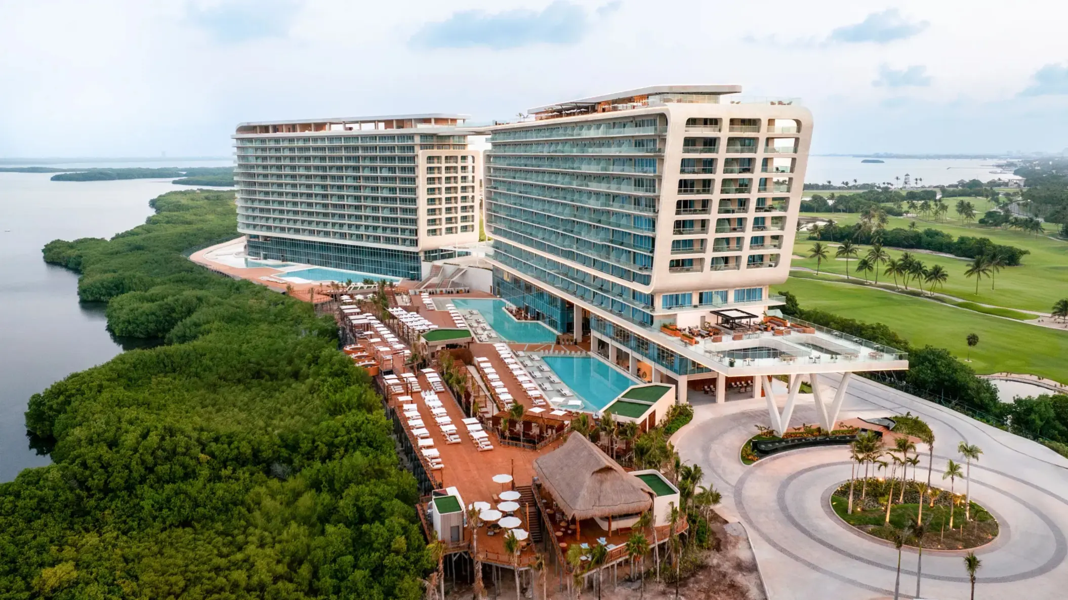 Wide aerial shot of beachfront resort with twin towers, rooftop pools, and lush mangroves all around.
