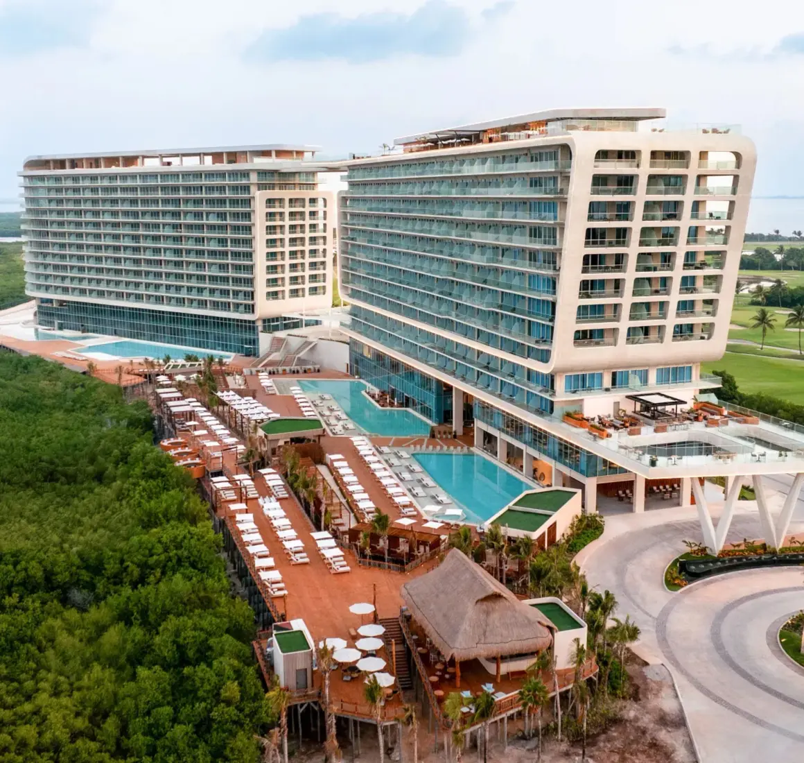 Wide aerial shot of beachfront resort with twin towers, rooftop pools, and lush mangroves all around.