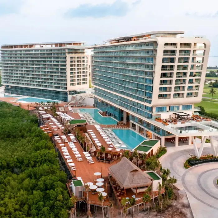 Wide aerial shot of beachfront resort with twin towers, rooftop pools, and lush mangroves all around.