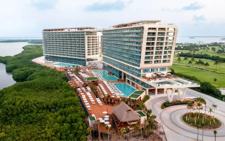 Wide aerial shot of beachfront resort with twin towers, rooftop pools, and lush mangroves all around.