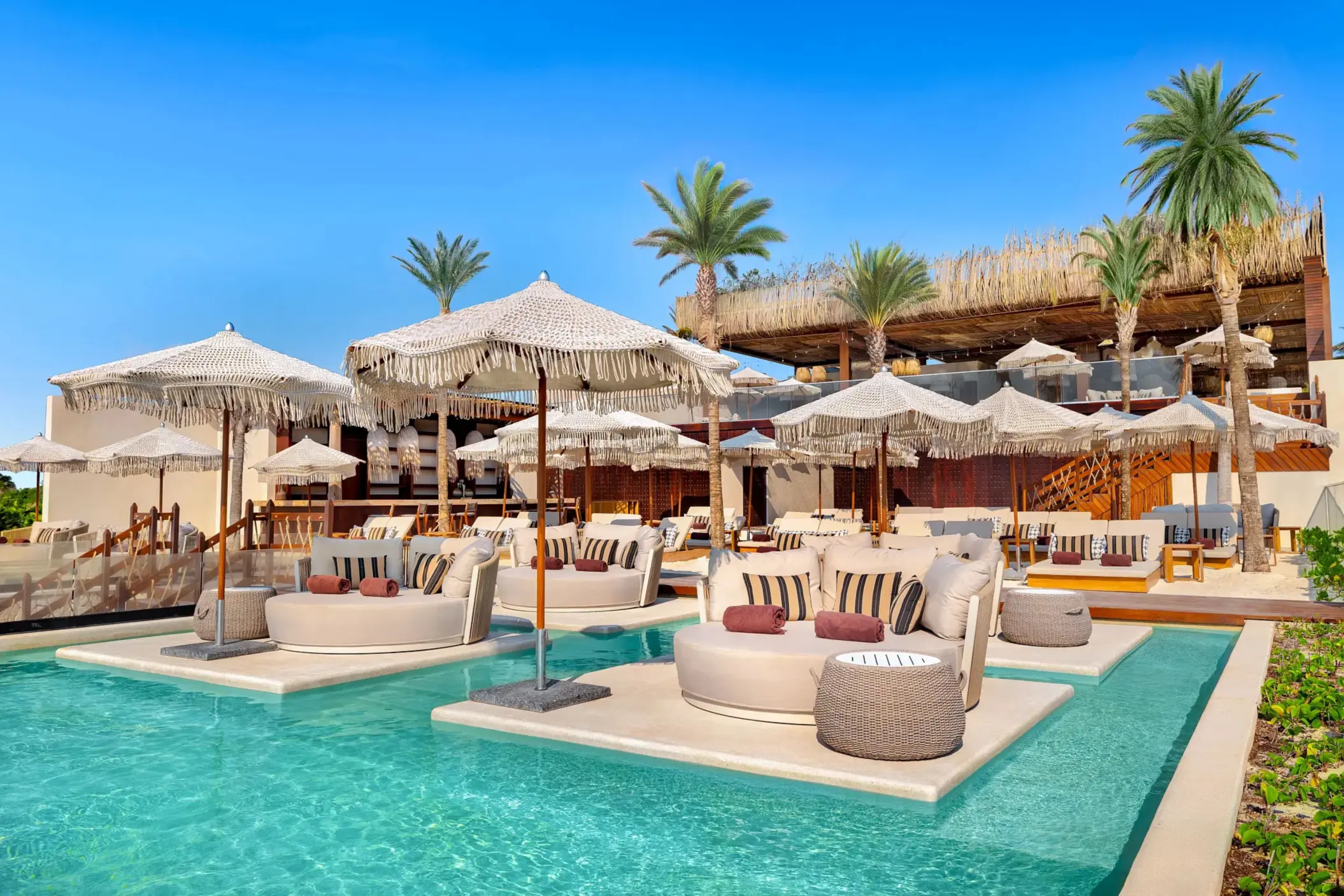 Shallow pool with in-water lounge beds, woven umbrellas, and cozy seating in a tropical beach club.