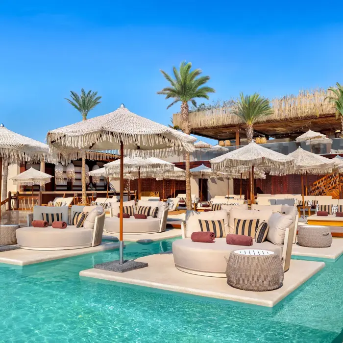 Shallow pool with in-water lounge beds, woven umbrellas, and cozy seating in a tropical beach club.