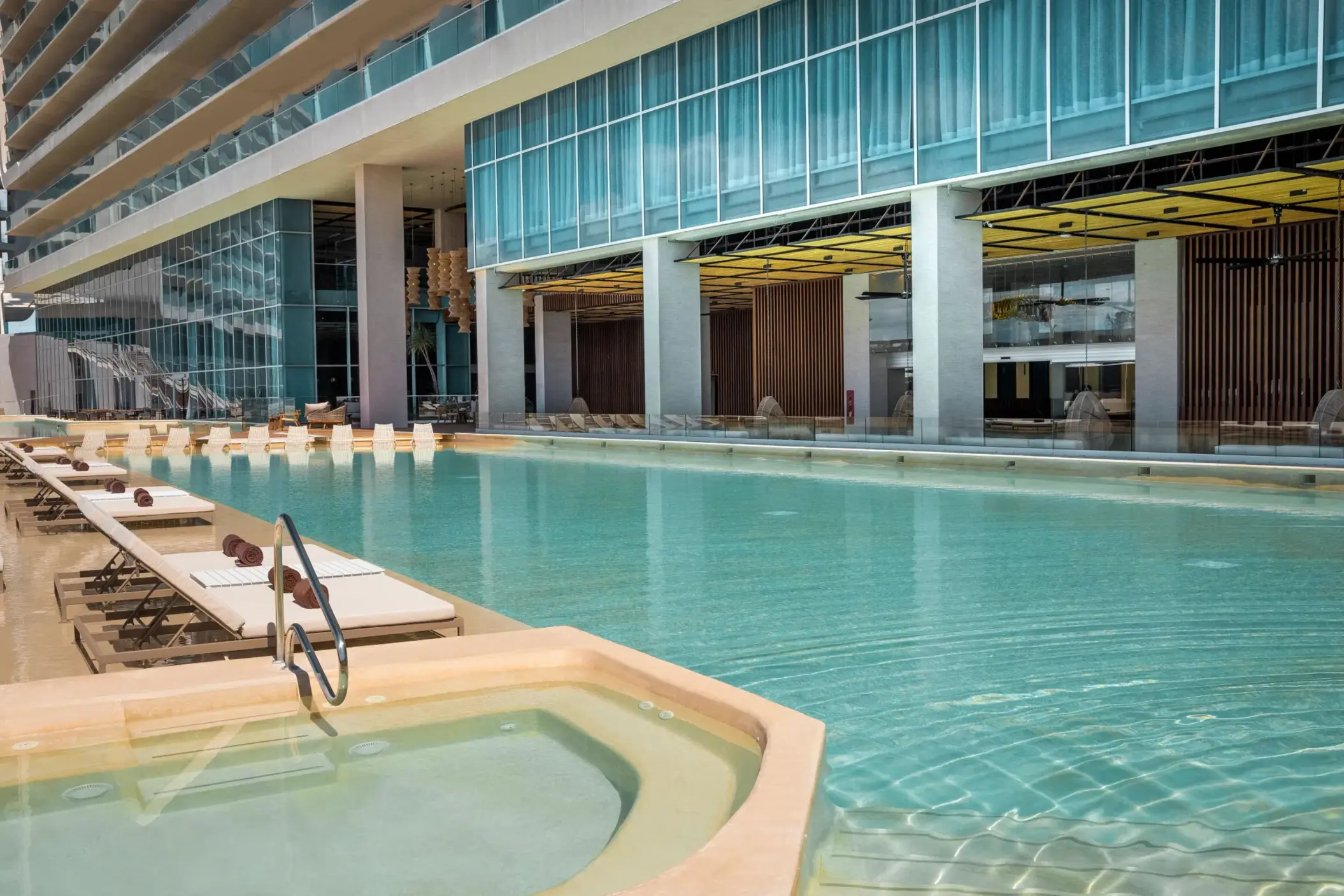 Long pool lined with white loungers, a hot tub, and modern resort exterior with open-air design.