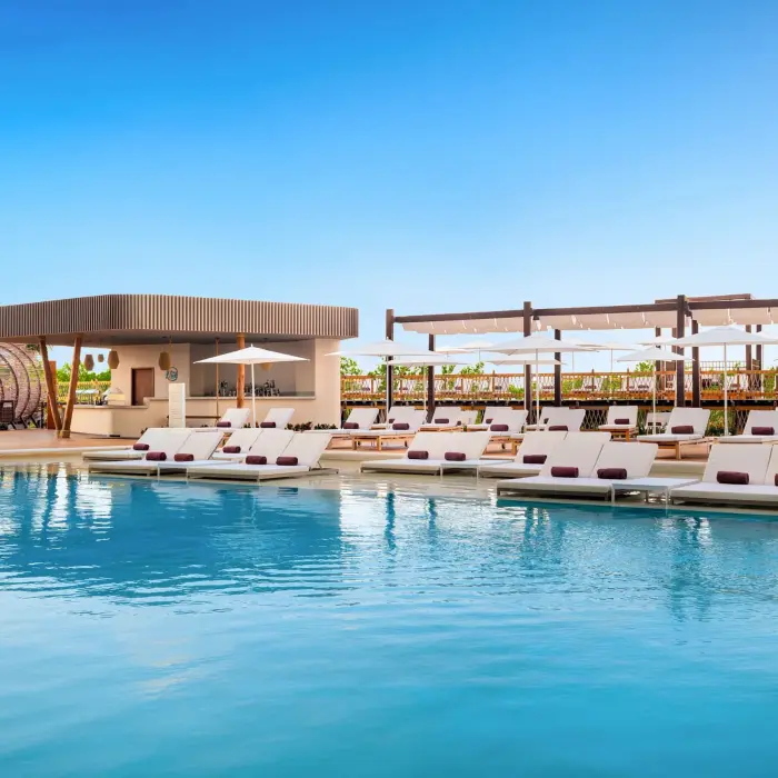 Main pool with modern loungers, white umbrellas, and a stylish poolside bar surrounded by tropical greenery.