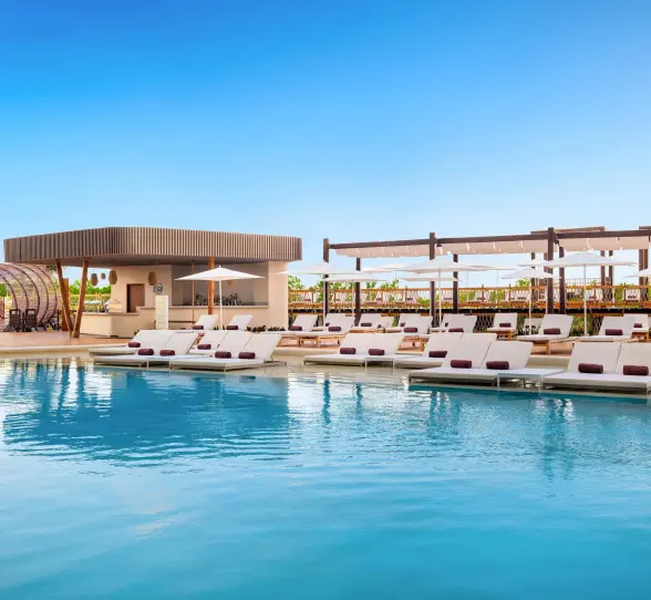 Main pool with modern loungers, white umbrellas, and a stylish poolside bar surrounded by tropical greenery.