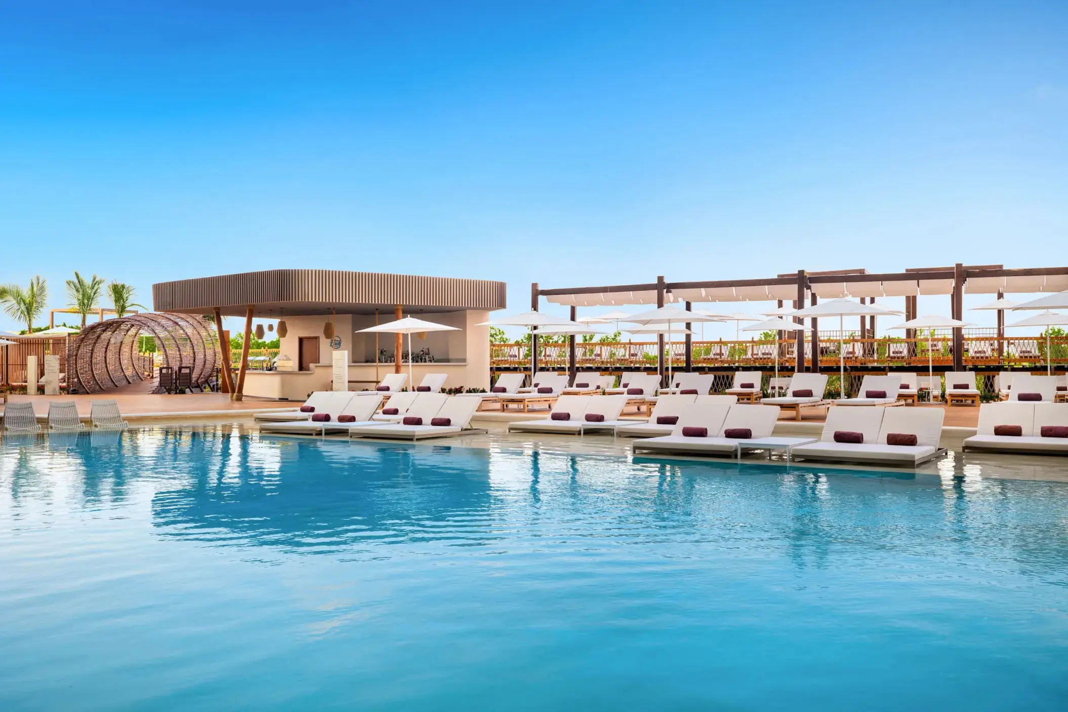 Main pool with modern loungers, white umbrellas, and a stylish poolside bar surrounded by tropical greenery.