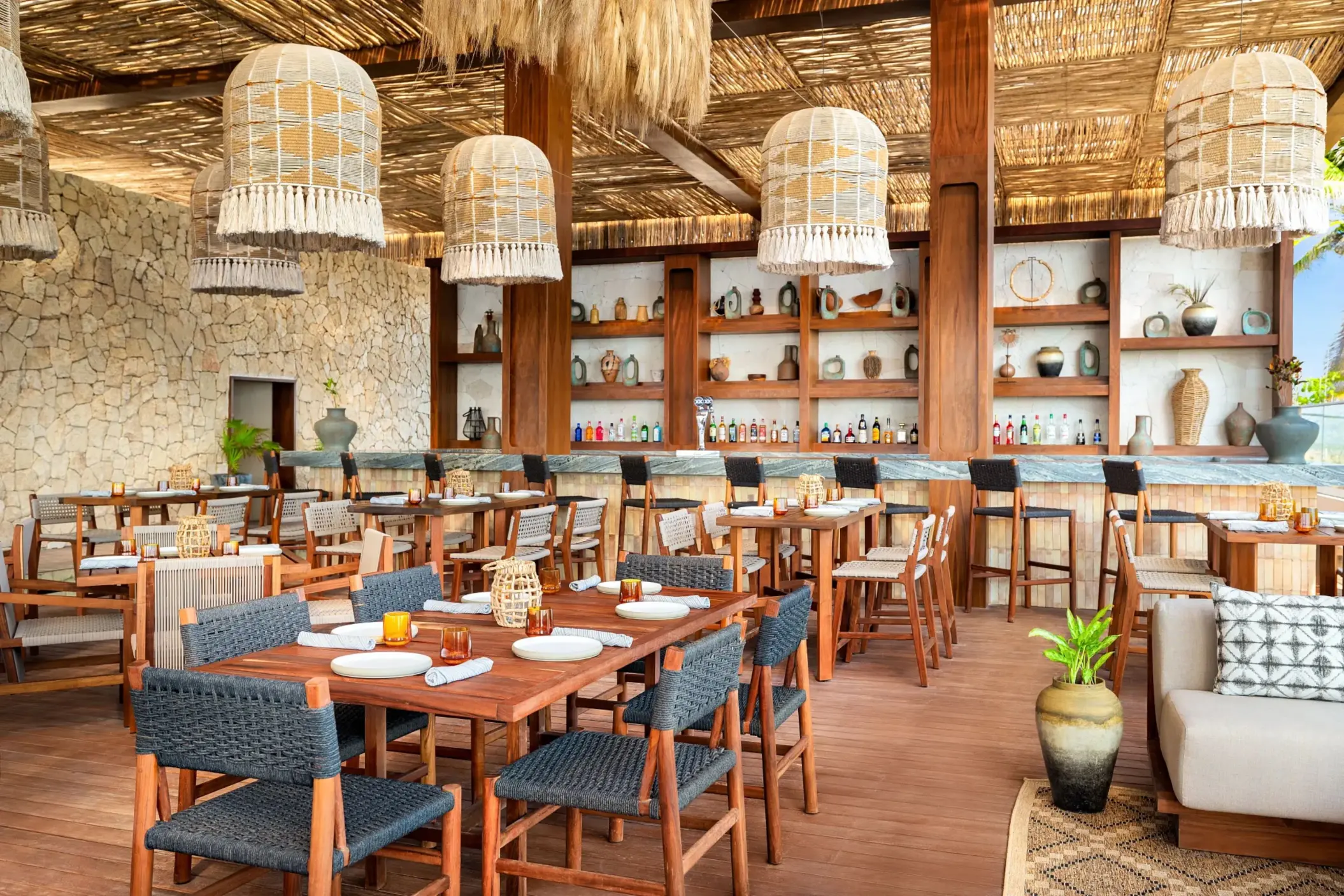 Rustic beach club restaurant with thatched ceiling, woven pendant lights, stone walls, and artisanal décor.