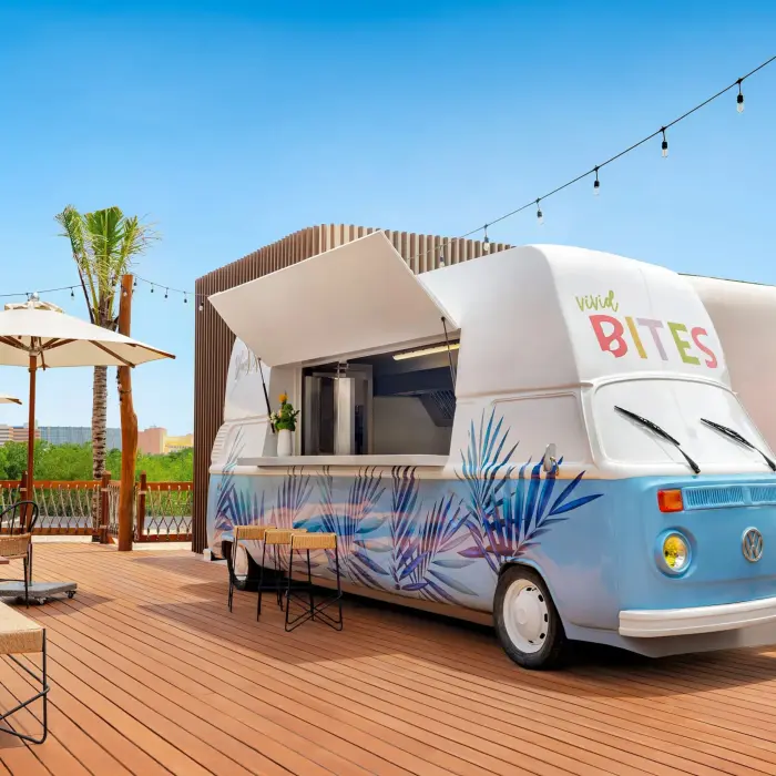 Colorful retro food truck labeled “Vivid Bites” on a sunny rooftop deck with string lights and palm trees.
