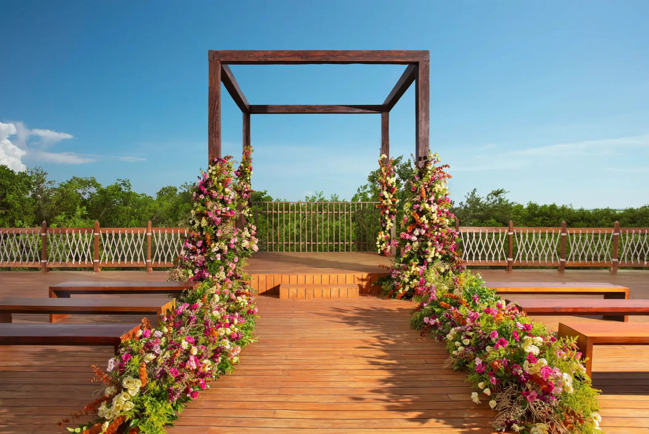 Outdoor wooden gazebo wedding setup with colorful floral aisle décor surrounded by tropical greenery and blue skies.