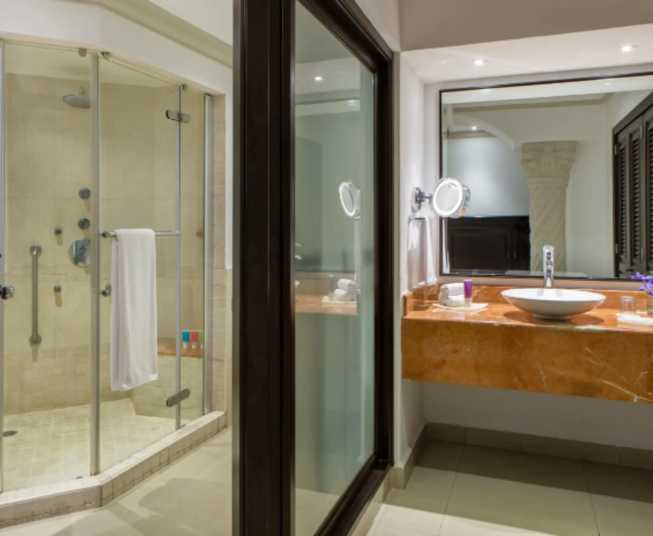 Modern resort bathroom with glass-enclosed shower, marble countertop, round sink, and large mirror near elegant lighting.