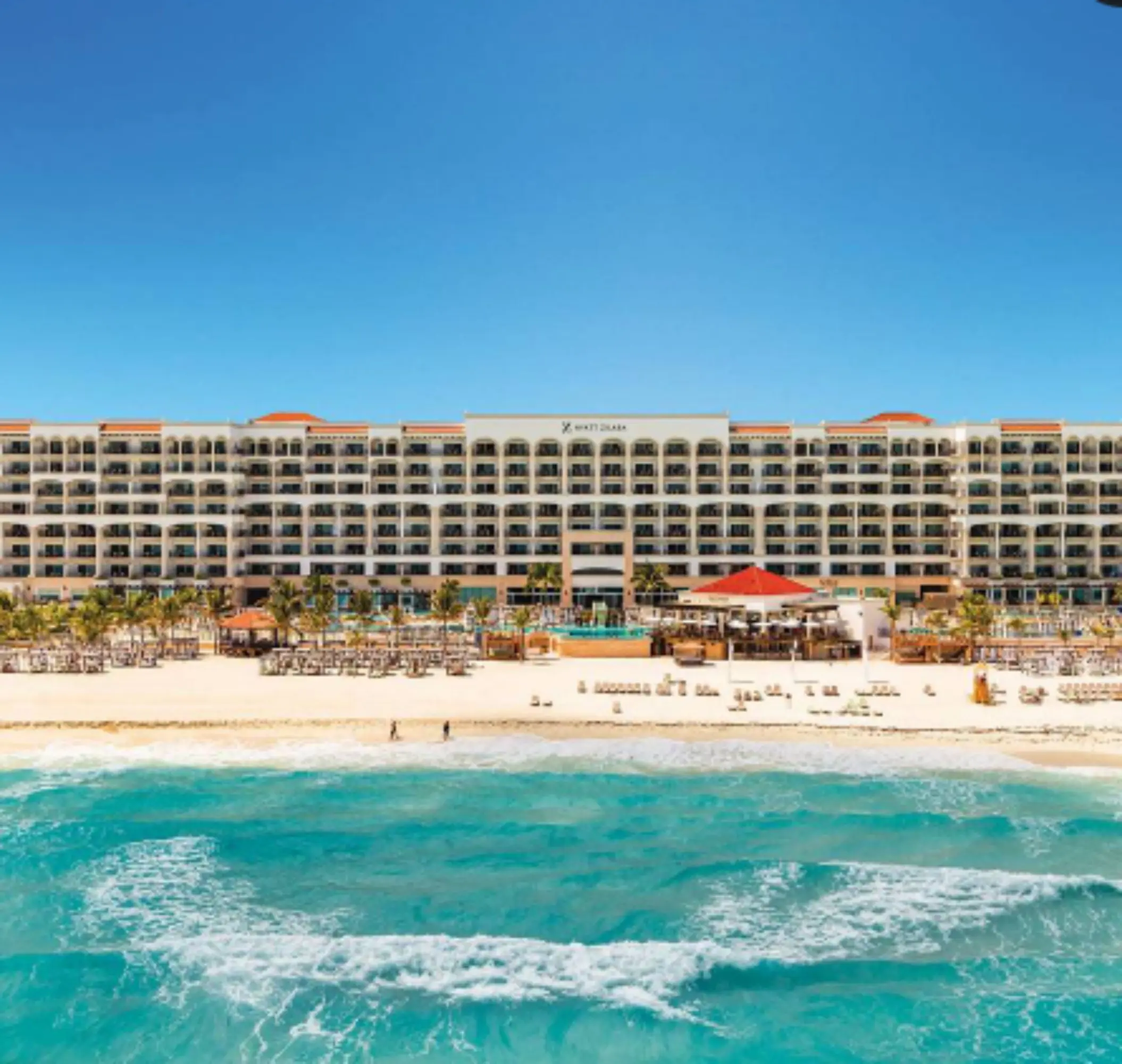 Oceanfront resort view showcasing balconies, beach loungers, and turquoise waves under a blue sky.