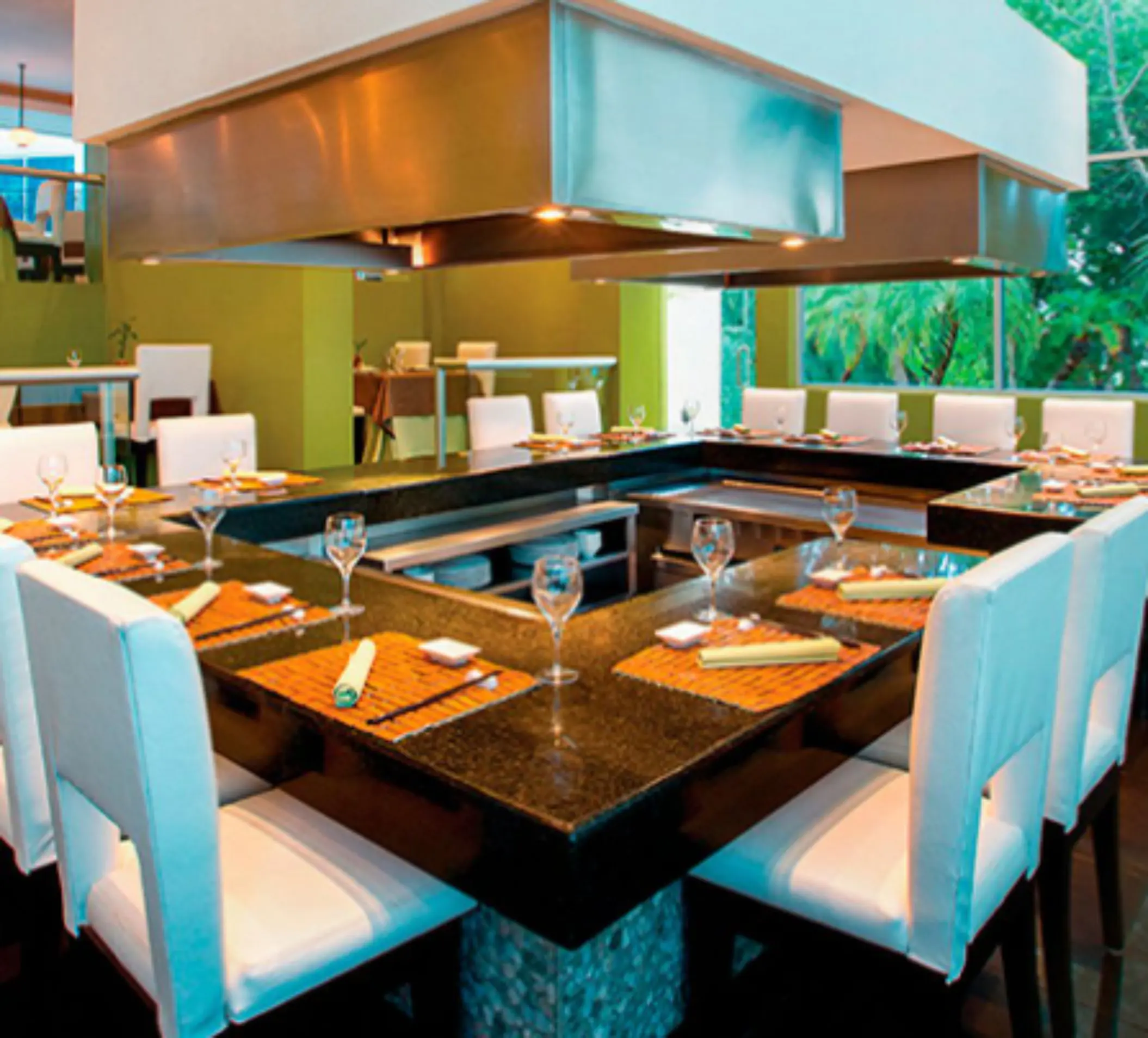 Elegant teppanyaki-style restaurant setup with white chairs and polished black countertops, surrounded by greenery.