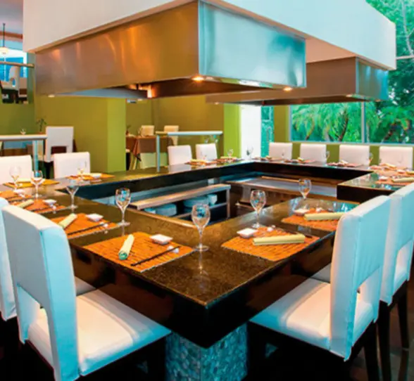 Elegant teppanyaki-style restaurant setup with white chairs and polished black countertops, surrounded by greenery.