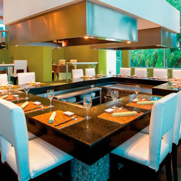 Elegant teppanyaki-style restaurant setup with white chairs and polished black countertops, surrounded by greenery.