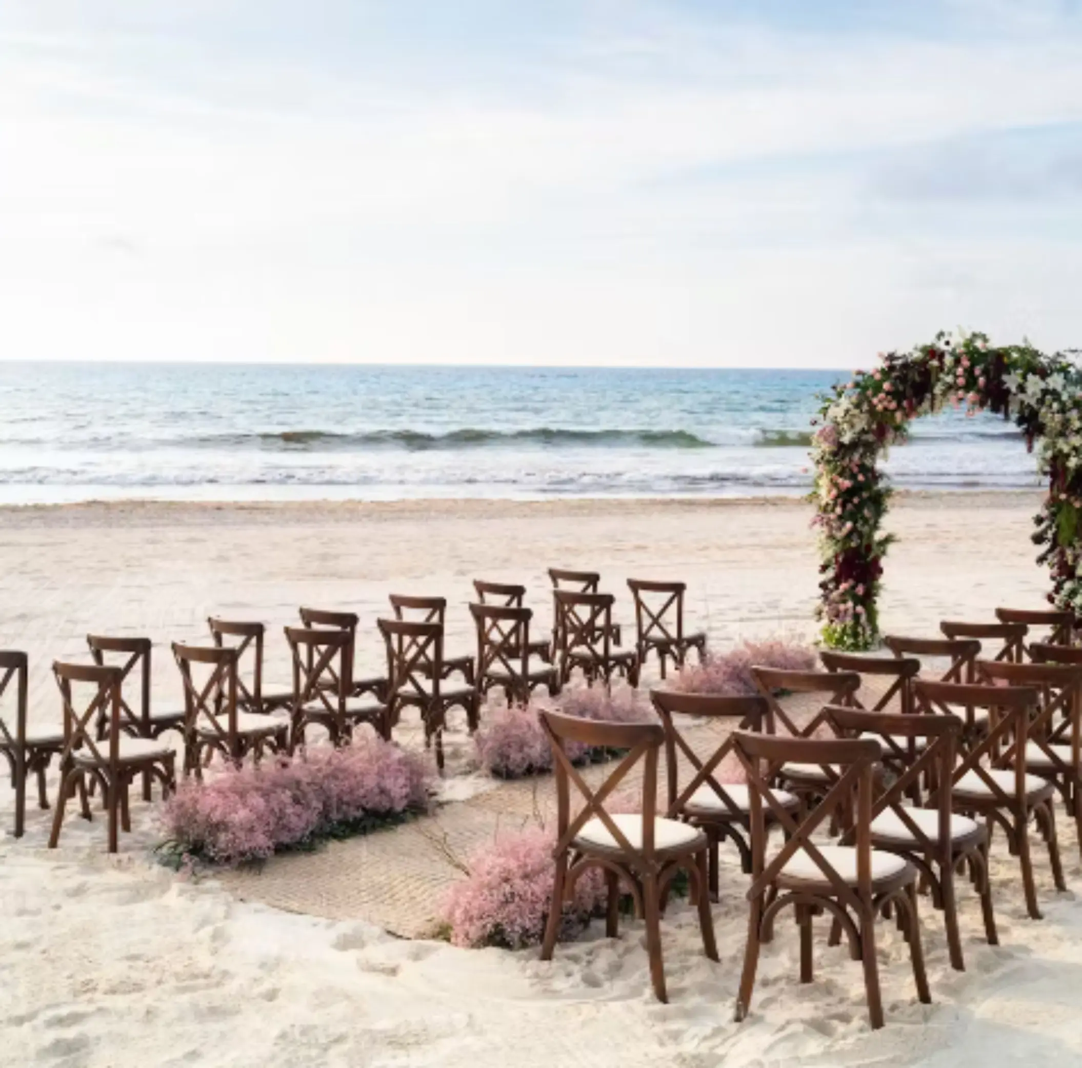 Beach wedding setup with wooden chairs, pink florals, and floral arch facing the ocean under a calm sky.