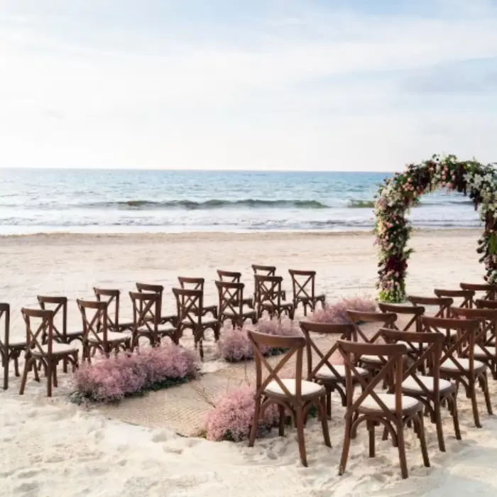 Beach wedding setup with wooden chairs, pink florals, and floral arch facing the ocean under a calm sky.