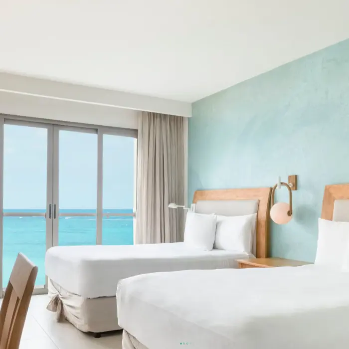 Bright guest room with two queen beds, oceanfront view, light wood furniture, and seafoam green accent wall.