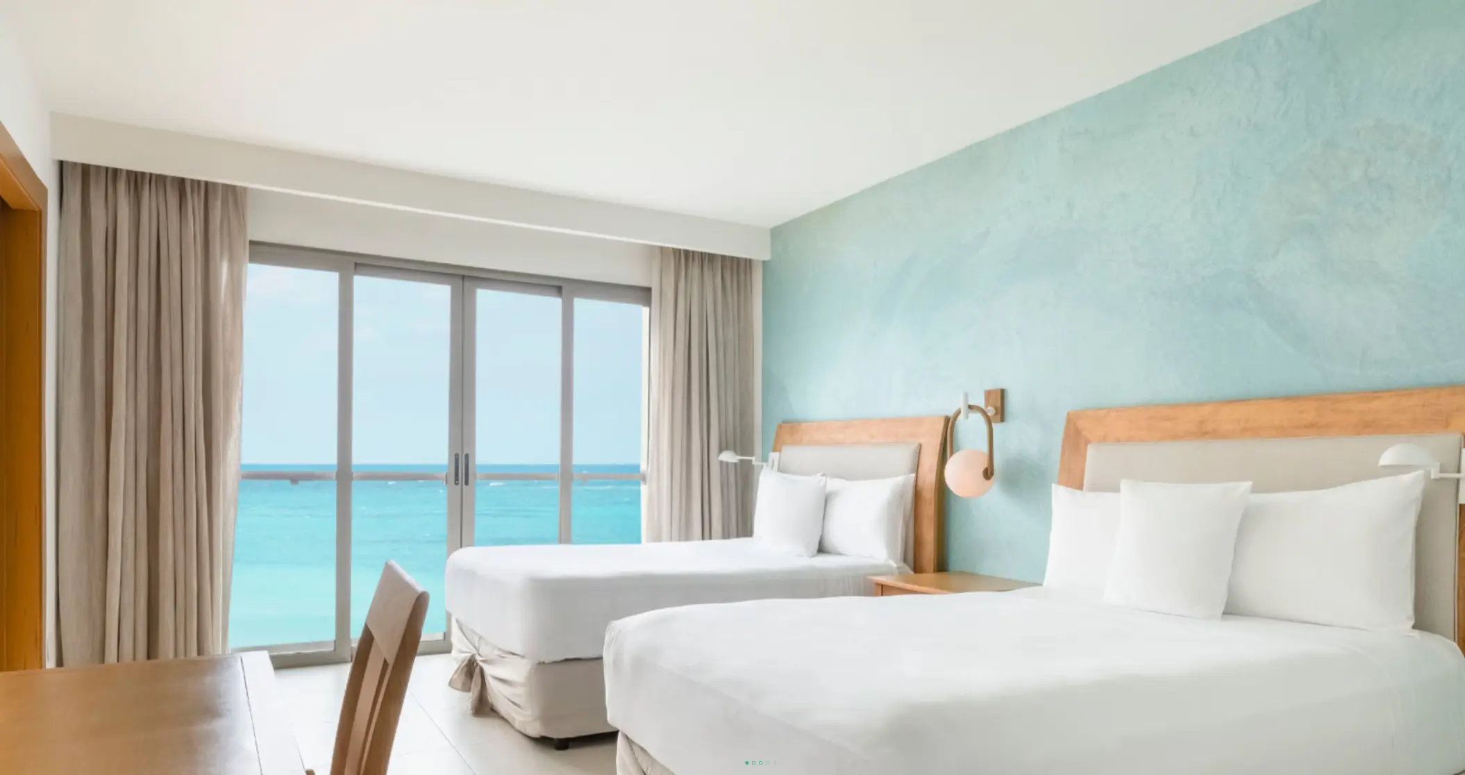 Bright guest room with two queen beds, oceanfront view, light wood furniture, and seafoam green accent wall.