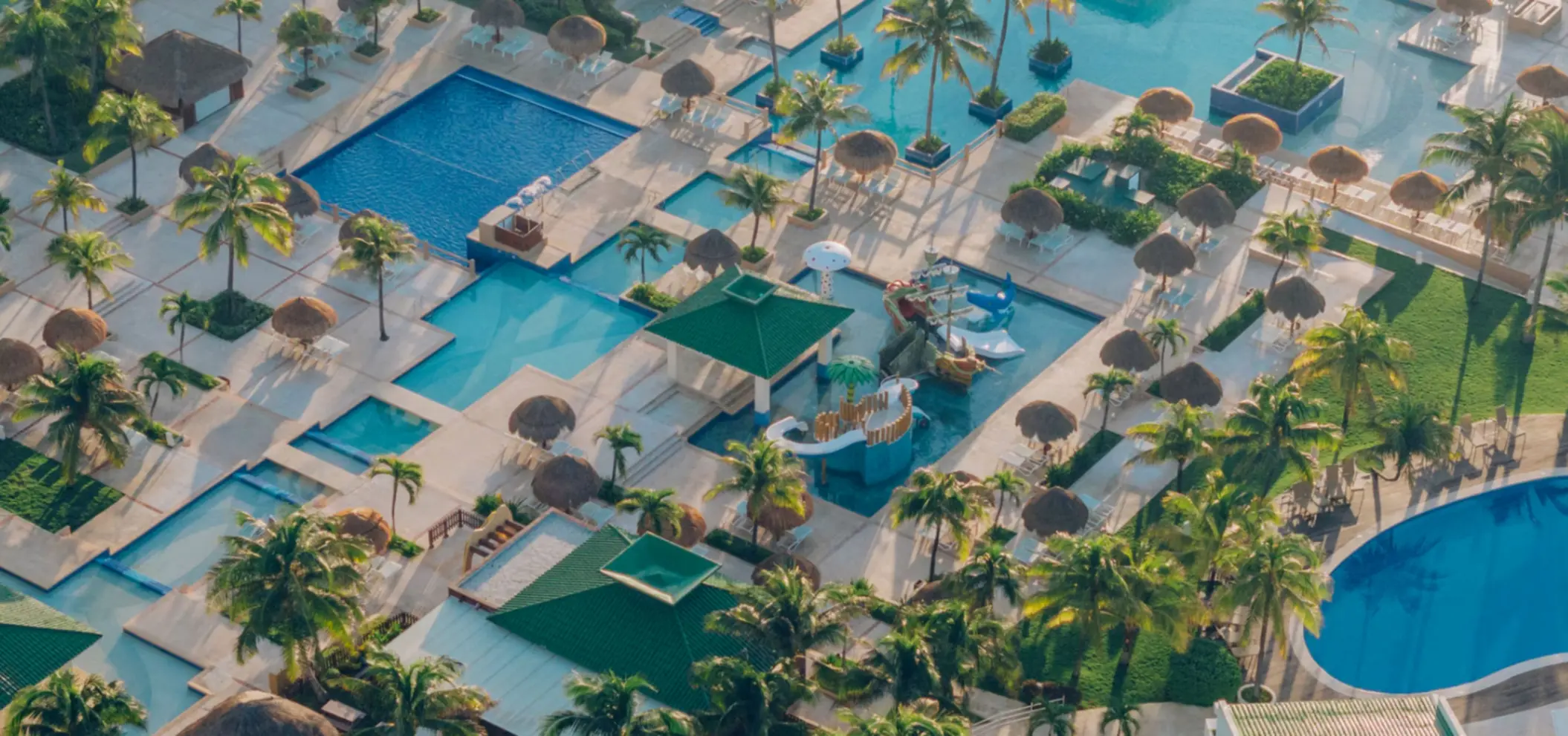 Aerial view of multi-pool complex with thatched-roof loungers, kids splash zone, and lush palm-filled landscape.