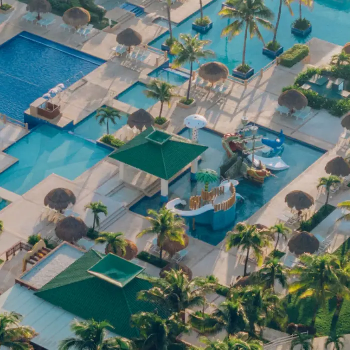 Aerial view of multi-pool complex with thatched-roof loungers, kids splash zone, and lush palm-filled landscape.