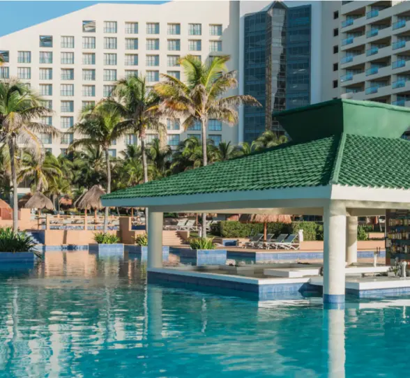 Poolside swim-up bar with green-tile roof surrounded by palm trees and resort buildings under clear blue skies.
