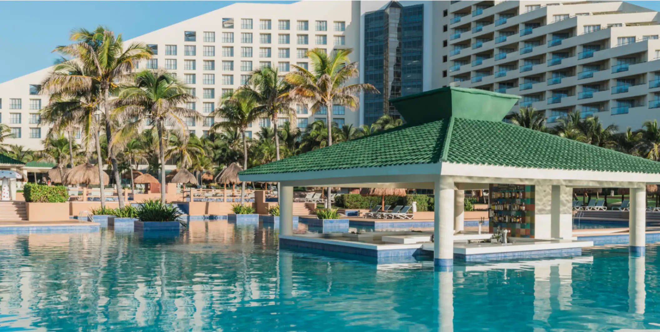 Poolside swim-up bar with green-tile roof surrounded by palm trees and resort buildings under clear blue skies.