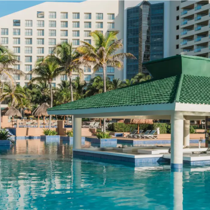 Poolside swim-up bar with green-tile roof surrounded by palm trees and resort buildings under clear blue skies.