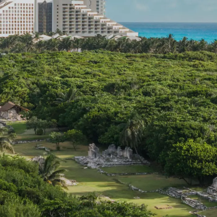 Aerial view of lush archaeological site surrounded by jungle, with stone ruins and thatched huts near the coastline.