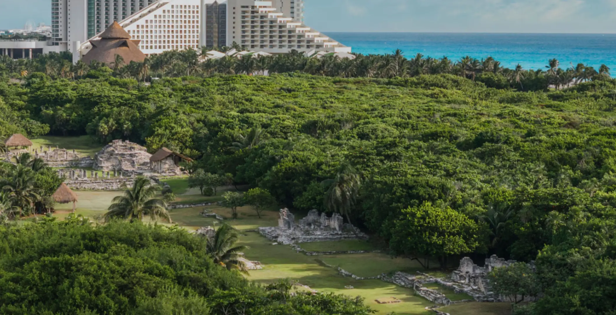 Aerial view of lush archaeological site surrounded by jungle, with stone ruins and thatched huts near the coastline.