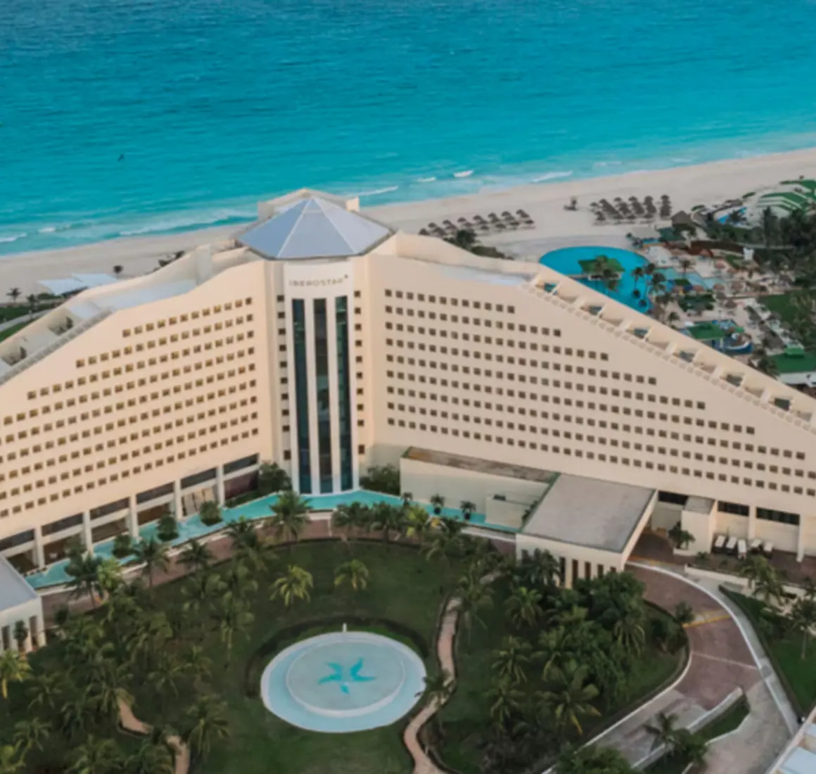 Panoramic aerial view of a beachfront resort with triangular hotel facade, pools, and turquoise Caribbean waters.