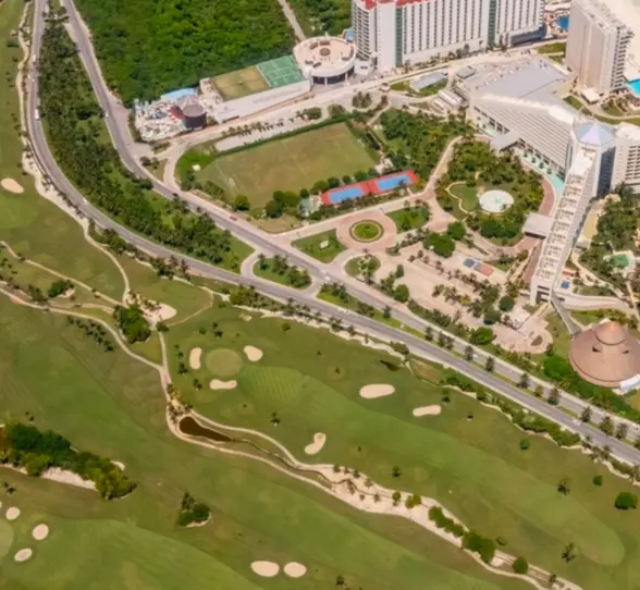 Aerial view of resort complex bordered by a green golf course, sandy beach, and bright blue ocean shoreline.