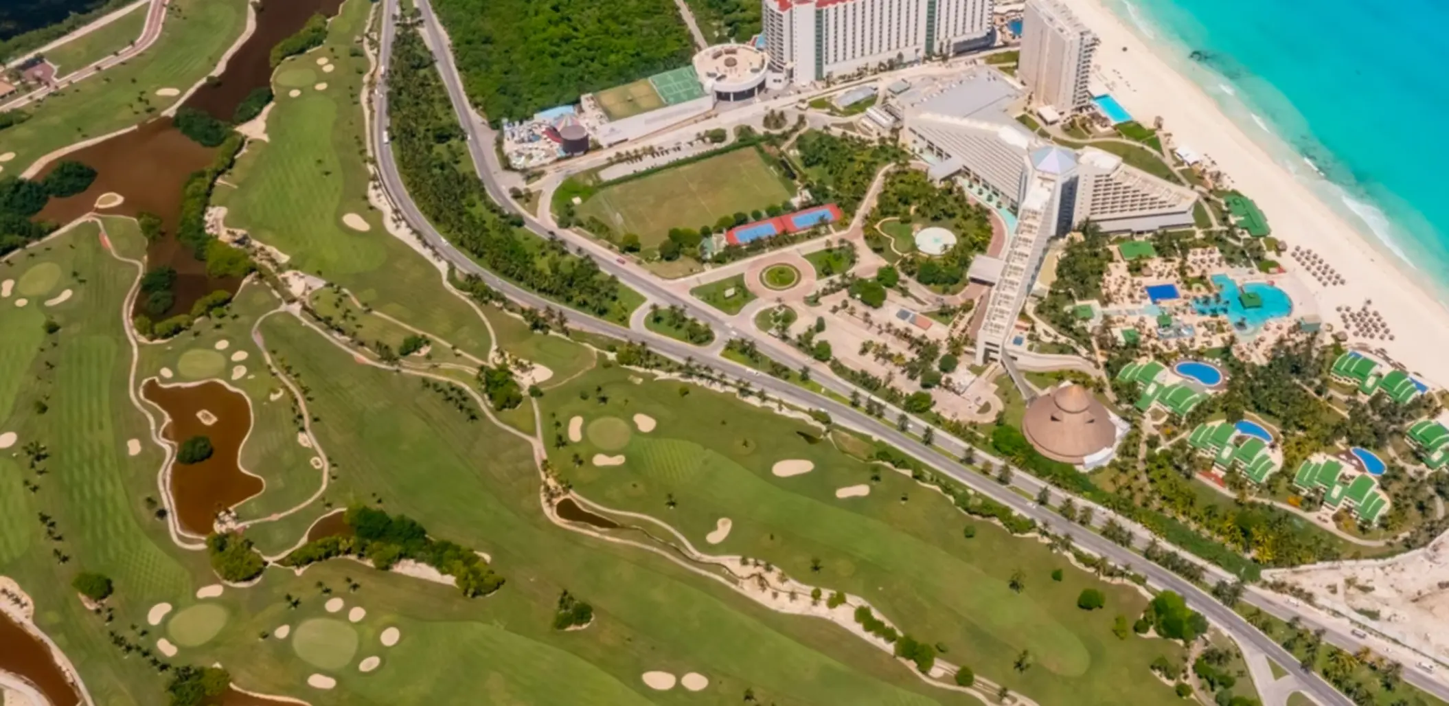 Aerial view of resort complex bordered by a green golf course, sandy beach, and bright blue ocean shoreline.