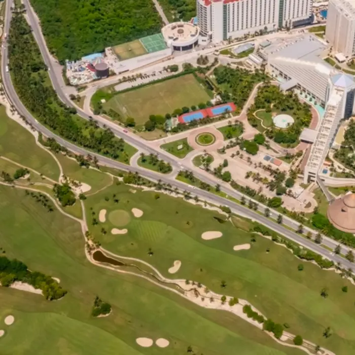 Aerial view of resort complex bordered by a green golf course, sandy beach, and bright blue ocean shoreline.