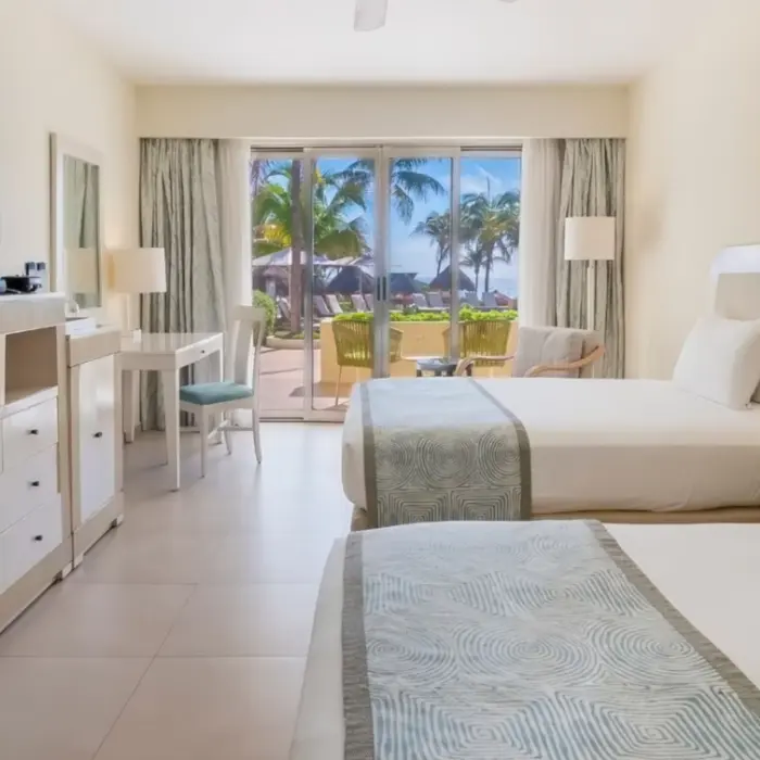 Bright guest room with two beds, a TV, writing desk, and ocean-facing terrace with palm trees and outdoor seating.