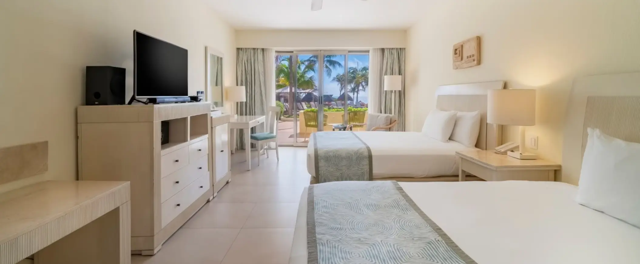 Bright guest room with two beds, a TV, writing desk, and ocean-facing terrace with palm trees and outdoor seating.