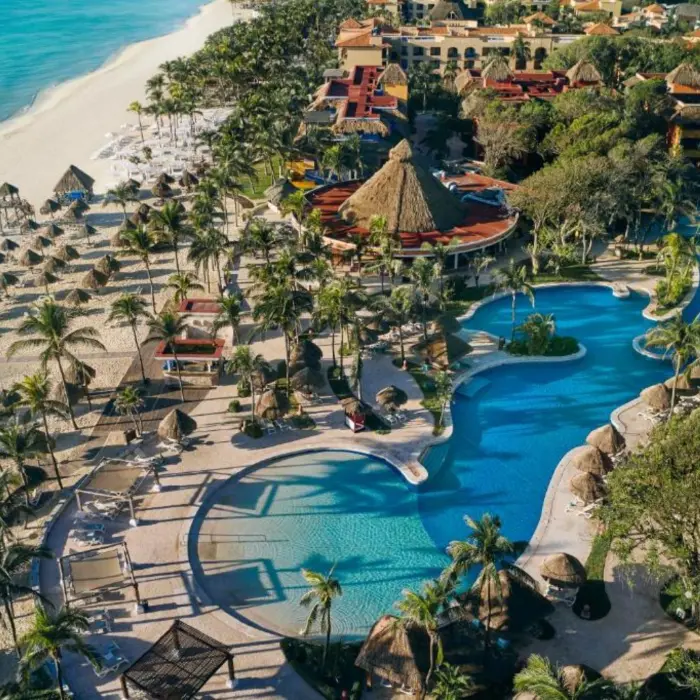 An aerial view of the hotel's pool area, palm trees, and beach with thatched umbrellas.