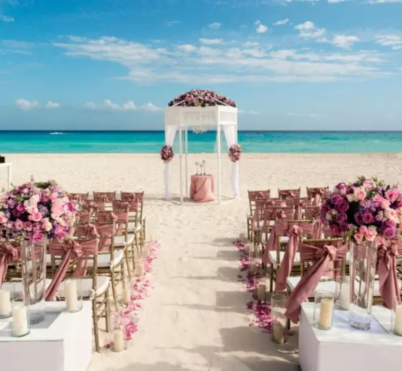 A beach wedding setup with a white arbor, rows of chairs, and pink flowers overlooking the sea.
