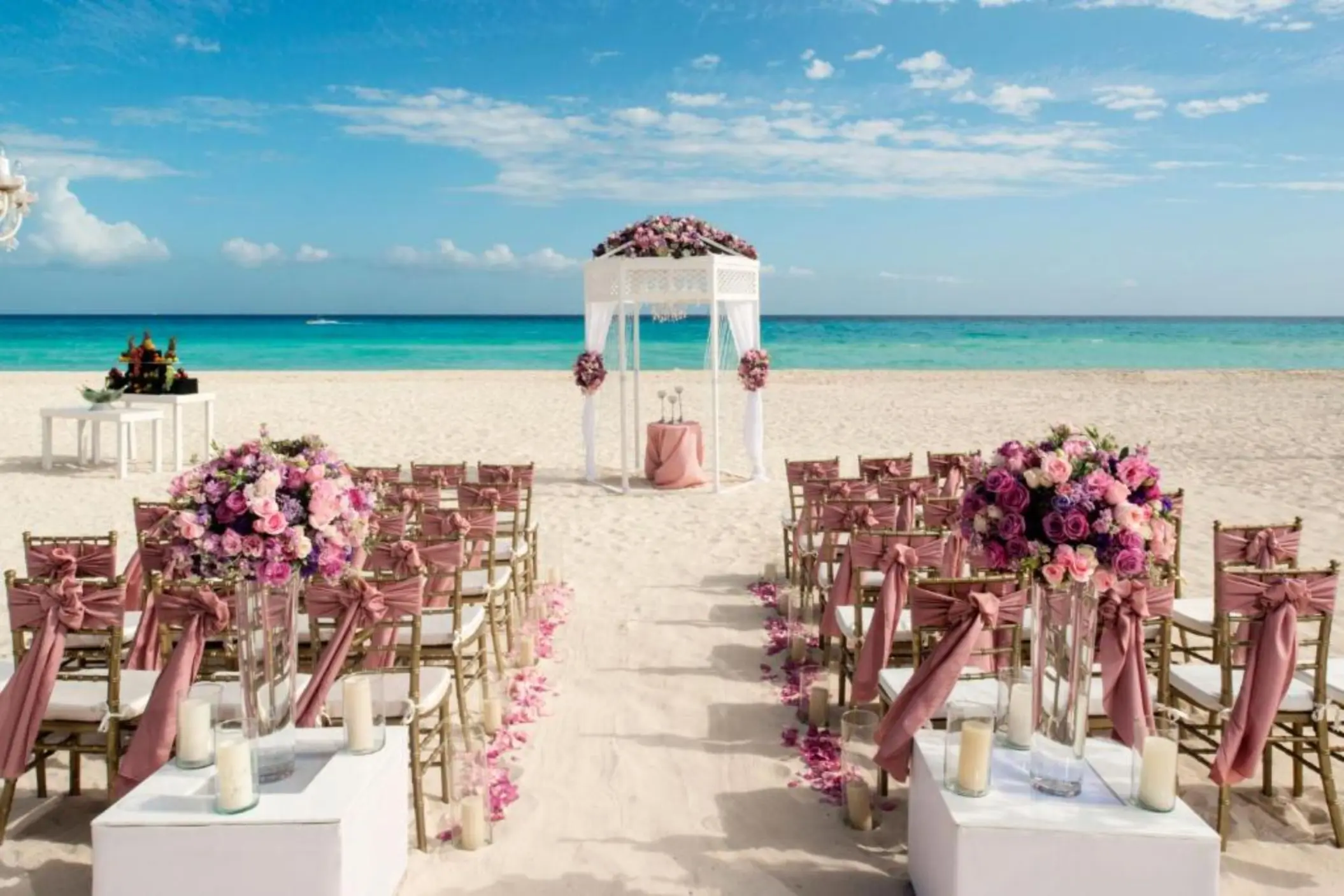 A beach wedding setup with a white arbor, rows of chairs, and pink flowers overlooking the sea.