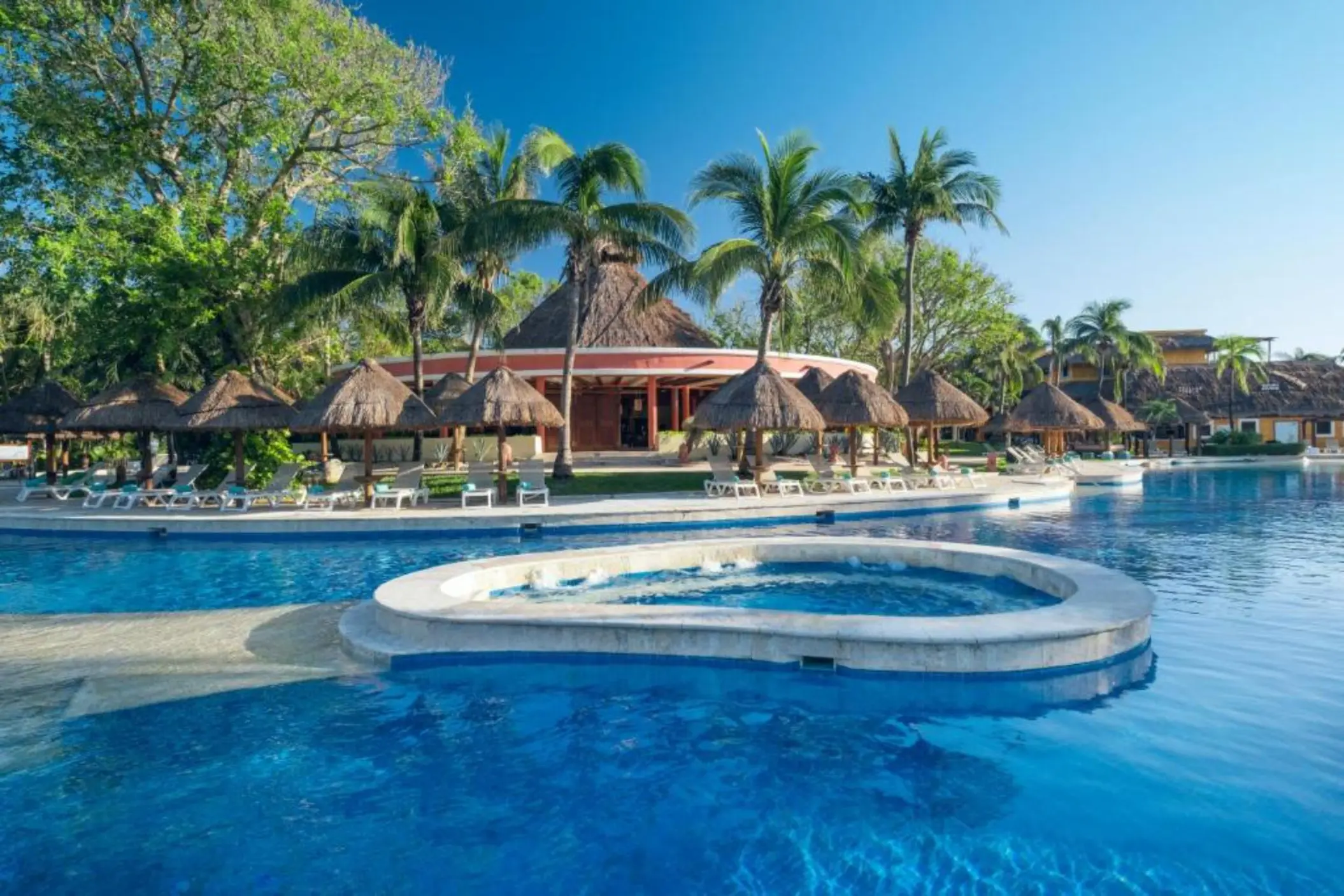 A large outdoor swimming pool with palm trees, lounge chairs, and a bar area.