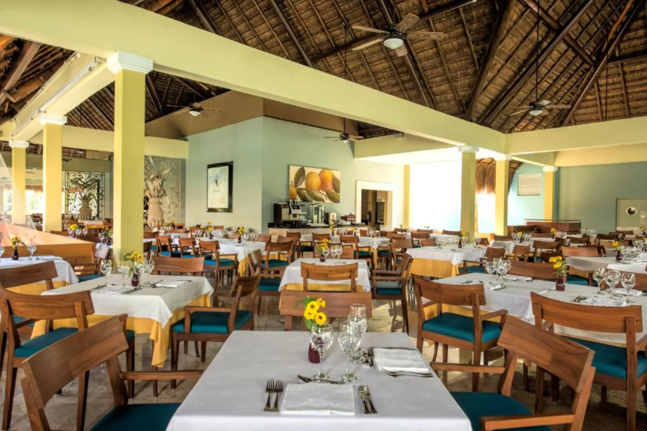 An empty restaurant with many tables and chairs set up, a thatched roof, and large windows.